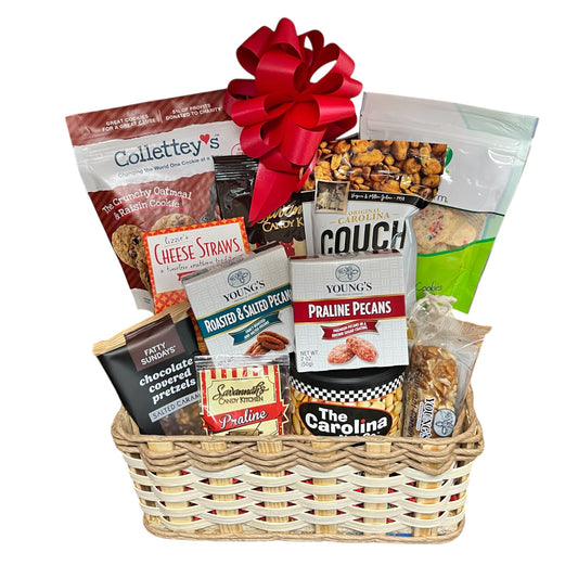 A wicker gift basket filled with assorted packaged snacks and cookies, topped with a large red bow. Visible snack packages include Colletty's, Young's, Carolina Couch, and Southern Comfort.